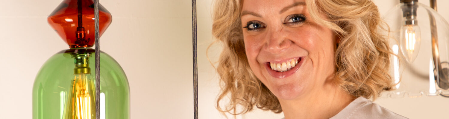 A smiling woman with blonde, wavy hair and blue eyes, wearing a white t-shirt. She is standing next to a modern light fixture that features multiple stacked, coloured glass shades, each containing an exposed filament bulb. The background is simple and well-lit, suggesting a showroom or interior setting.