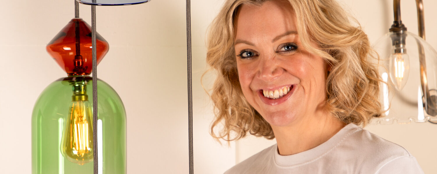 A smiling woman with blonde, wavy hair and blue eyes, wearing a white t-shirt. She is standing next to a modern light fixture that features multiple stacked, coloured glass shades, each containing an exposed filament bulb. The background is simple and well-lit, suggesting a showroom or interior setting.
