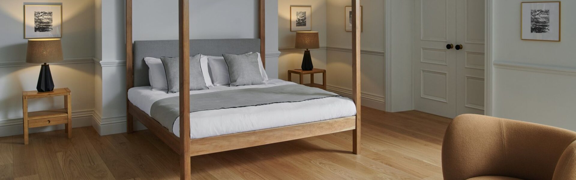 A bright, contemporary bedroom with light hardwood floors and pale grey walls. The centrepiece is a wooden four-poster bed with a grey upholstered headboard, white linens, and a grey throw blanket, accented by several pillows in white and light grey.