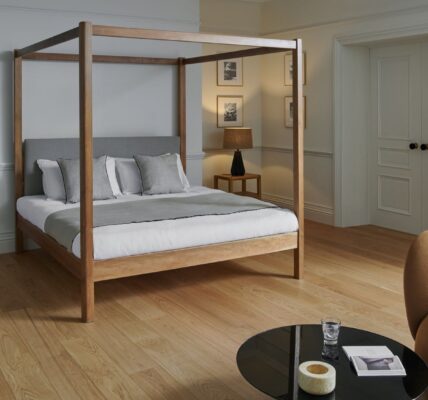 A bright, contemporary bedroom with light hardwood floors and pale grey walls. The centrepiece is a wooden four-poster bed with a grey upholstered headboard, white linens, and a grey throw blanket, accented by several pillows in white and light grey.