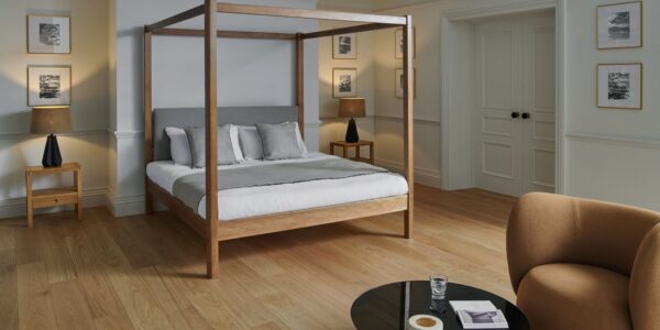 A bright, contemporary bedroom with light hardwood floors and pale grey walls. The centrepiece is a wooden four-poster bed with a grey upholstered headboard, white linens, and a grey throw blanket, accented by several pillows in white and light grey.