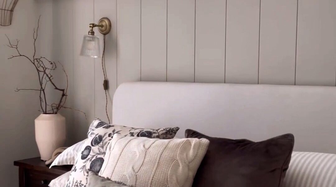 The wall features vertical V-groove panelling painted a light, neutral grey/taupe colour. The panelling extends upwards, perhaps to meet a picture rail or just below the ceiling, creating a backdrop for the bed.