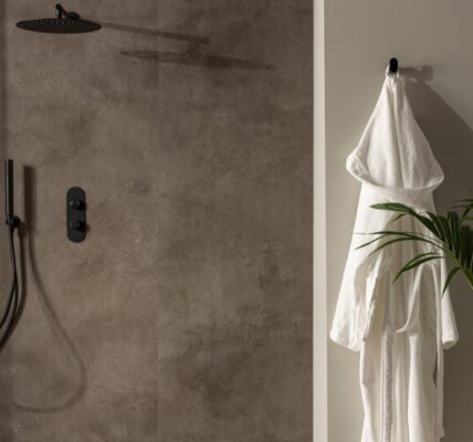 A shower wall clad in a dark, mottled, stone-effect panel. It has a warm, earthy brown/grey tone with slight texture variations. The shower fixtures—including the rainfall shower head, the handheld shower wand, and the control panel—are all in a matte black finish, providing a stark, contemporary contrast against the stone panel.