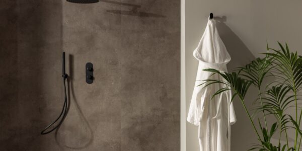 A shower wall clad in a dark, mottled, stone-effect panel. It has a warm, earthy brown/grey tone with slight texture variations. The shower fixtures—including the rainfall shower head, the handheld shower wand, and the control panel—are all in a matte black finish, providing a stark, contemporary contrast against the stone panel.