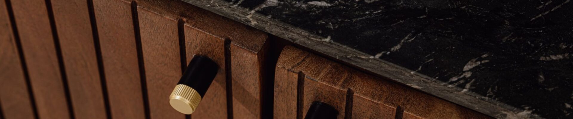 Close-up of a fluted wooden cabinet with two dual-tone cylindrical pulls in matte black and knurled gold, topped with a black marble counter.