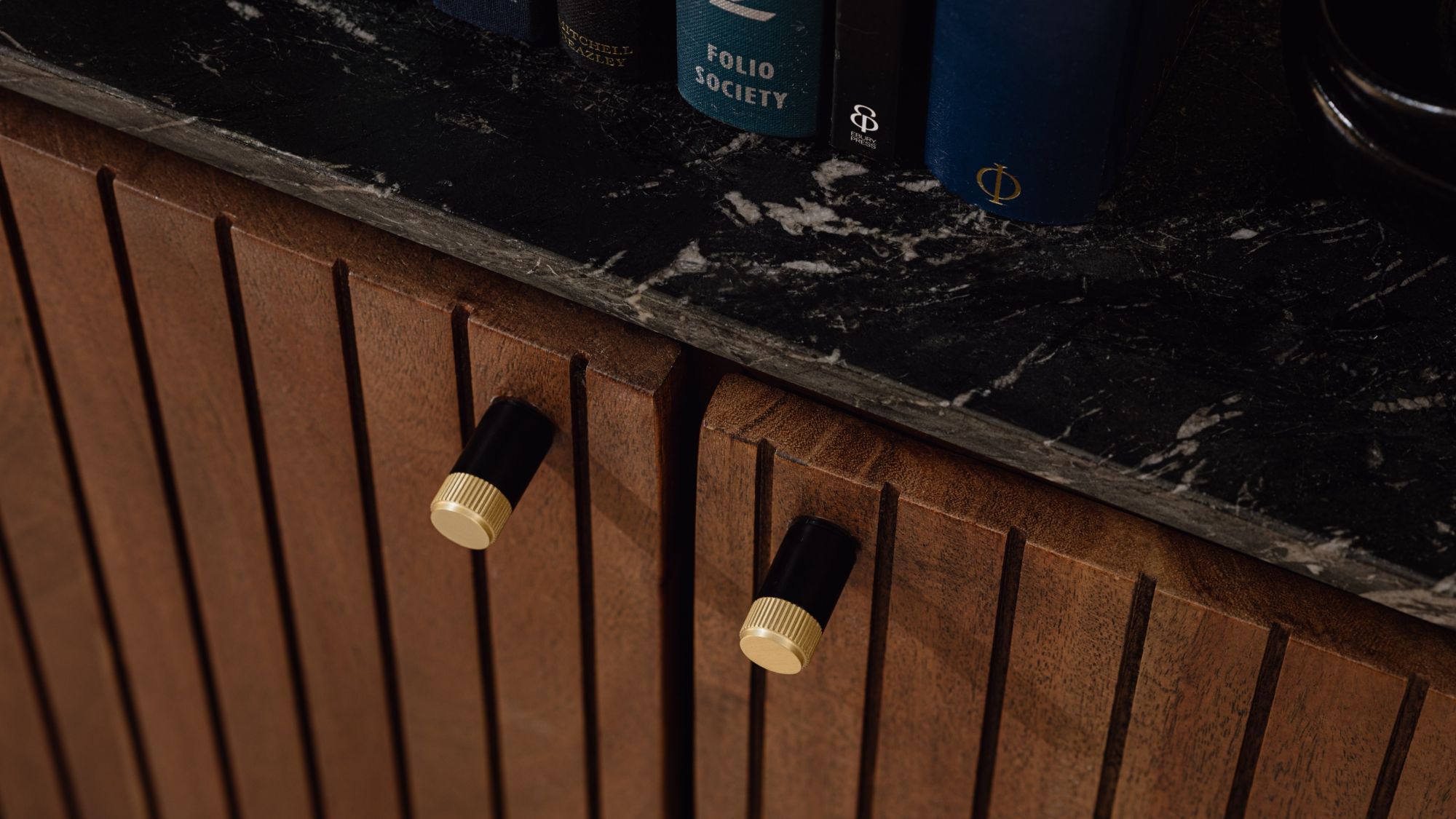 Close-up of a fluted wooden cabinet with two dual-tone cylindrical pulls in matte black and knurled gold, topped with a black marble counter.