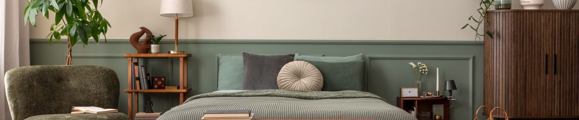 A cosy, biophilic-style bedroom featuring a bed with sage green linens and a textured moss-green throw. The wall behind the bed is split into a warm oatmeal top and muted green wood panelling on the bottom.