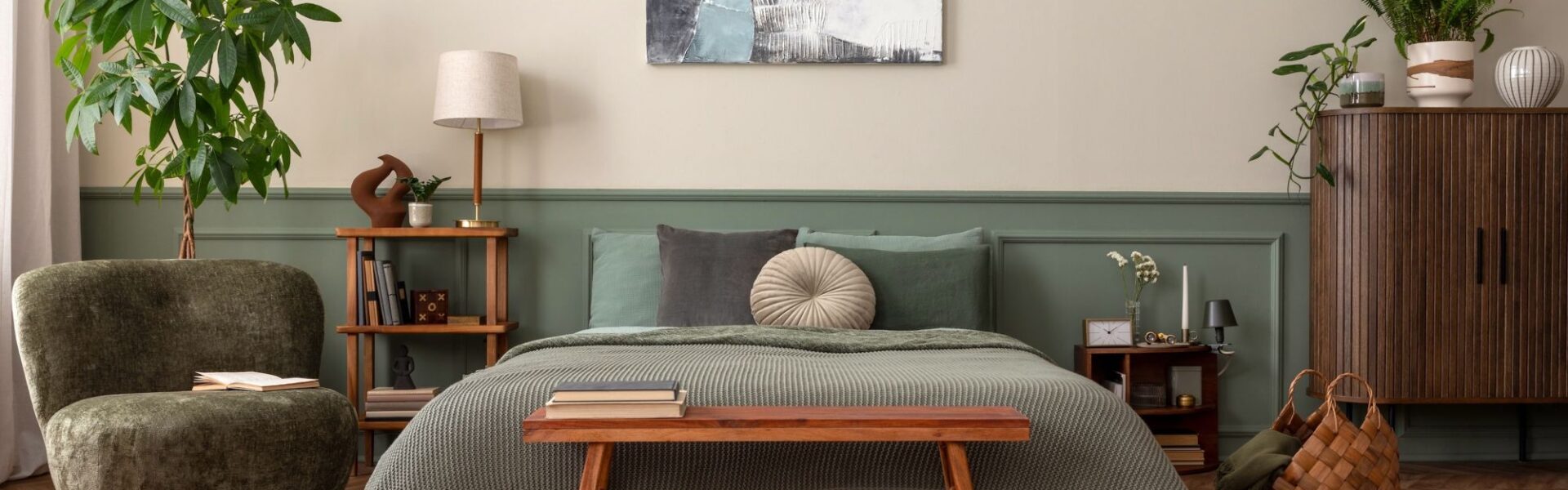 A cosy, biophilic-style bedroom featuring a bed with sage green linens and a textured moss-green throw. The wall behind the bed is split into a warm oatmeal top and muted green wood panelling on the bottom.