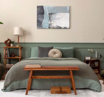 A cosy, biophilic-style bedroom featuring a bed with sage green linens and a textured moss-green throw. The wall behind the bed is split into a warm oatmeal top and muted green wood panelling on the bottom.