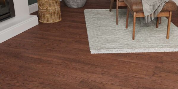 A cosy living room setting featuring Lusso Modena Luxe Oakmere engineered oak flooring in a rich, dark walnut finish. The dark wood planks are paired with a white fireplace hearth, a woven log basket, and a light grey patterned rug under mid-century modern furniture.