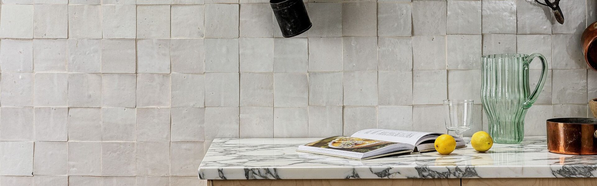 A kitchen interior featuring a handmade white Zellige tile backsplash with a textured, uneven surface. A marble countertop sits atop light wood cabinetry, holding an open cookbook, two lemons, and a green fluted glass pitcher. Above, a brass rail hangs against the tile with kitchen tools and a small black cup.
