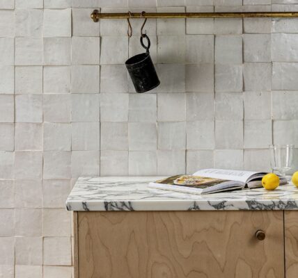 A kitchen interior featuring a handmade white Zellige tile backsplash with a textured, uneven surface. A marble countertop sits atop light wood cabinetry, holding an open cookbook, two lemons, and a green fluted glass pitcher. Above, a brass rail hangs against the tile with kitchen tools and a small black cup.