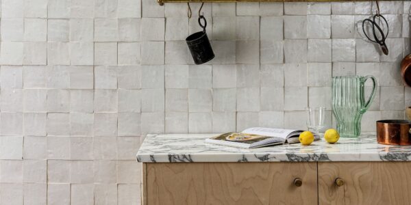 A kitchen interior featuring a handmade white Zellige tile backsplash with a textured, uneven surface. A marble countertop sits atop light wood cabinetry, holding an open cookbook, two lemons, and a green fluted glass pitcher. Above, a brass rail hangs against the tile with kitchen tools and a small black cup.