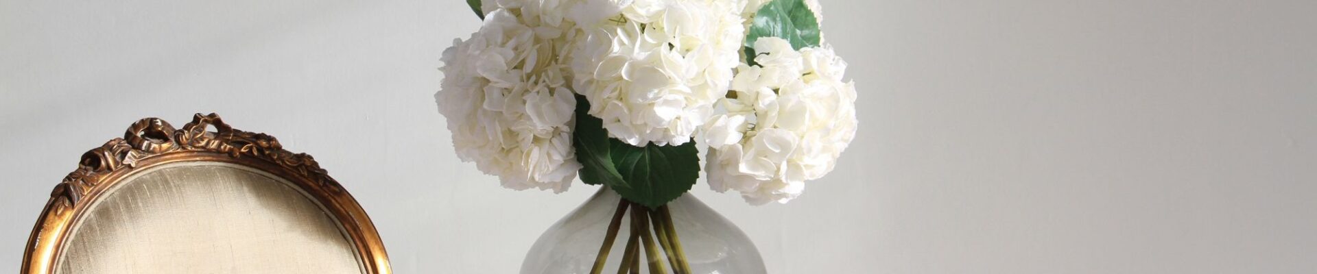 : An elegant indoor setup featuring a bouquet of artificial white hydrangeas in a round, smoky-grey glass vase. The vase sits on a small, gold-finished side table with a circular glass top and ornate geometric detailing.
