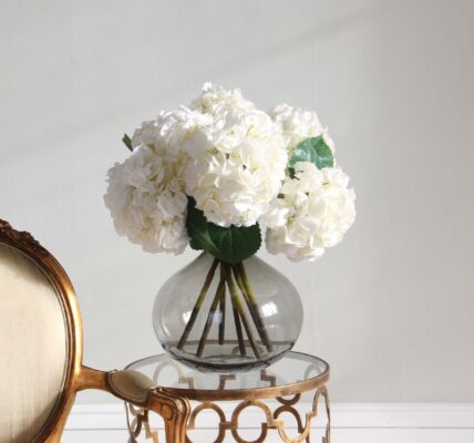 : An elegant indoor setup featuring a bouquet of artificial white hydrangeas in a round, smoky-grey glass vase. The vase sits on a small, gold-finished side table with a circular glass top and ornate geometric detailing.