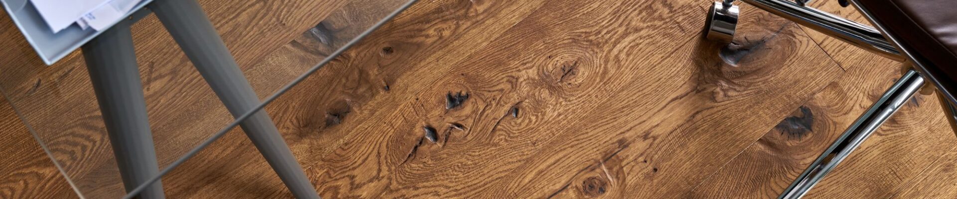 A top-down view of Berkeley Cathedral Oak engineered wood flooring featuring deep grain patterns and rustic knots, partially covered by a glass desk and a leather office chair.