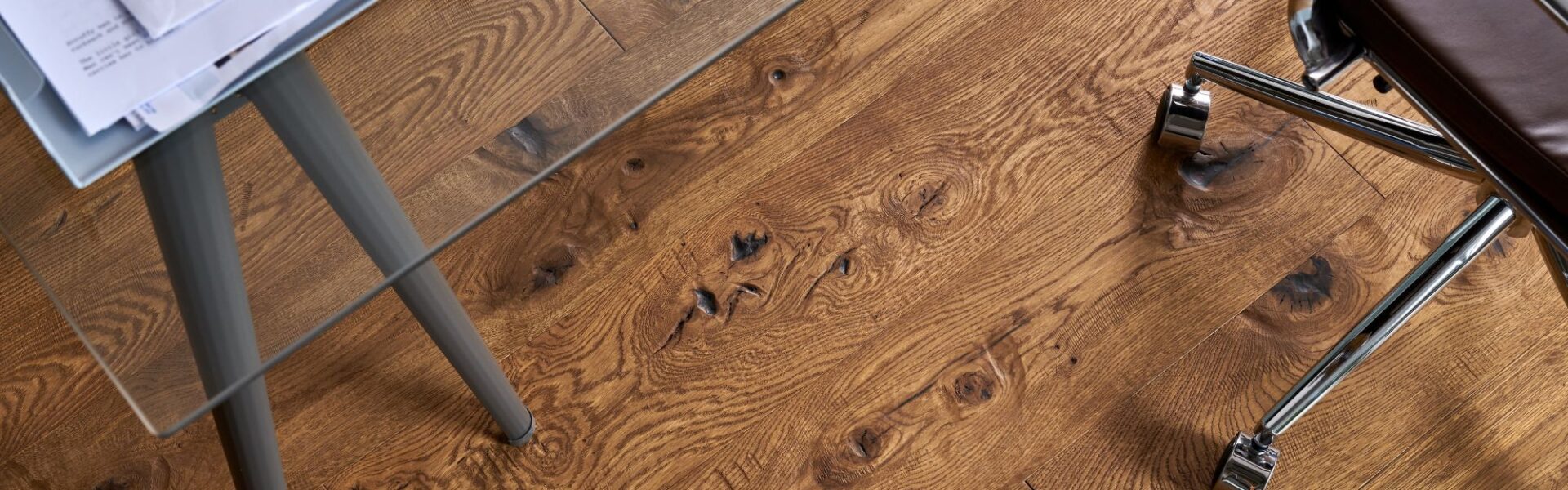 A top-down view of Berkeley Cathedral Oak engineered wood flooring featuring deep grain patterns and rustic knots, partially covered by a glass desk and a leather office chair.