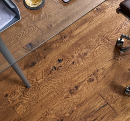 A top-down view of Berkeley Cathedral Oak engineered wood flooring featuring deep grain patterns and rustic knots, partially covered by a glass desk and a leather office chair.