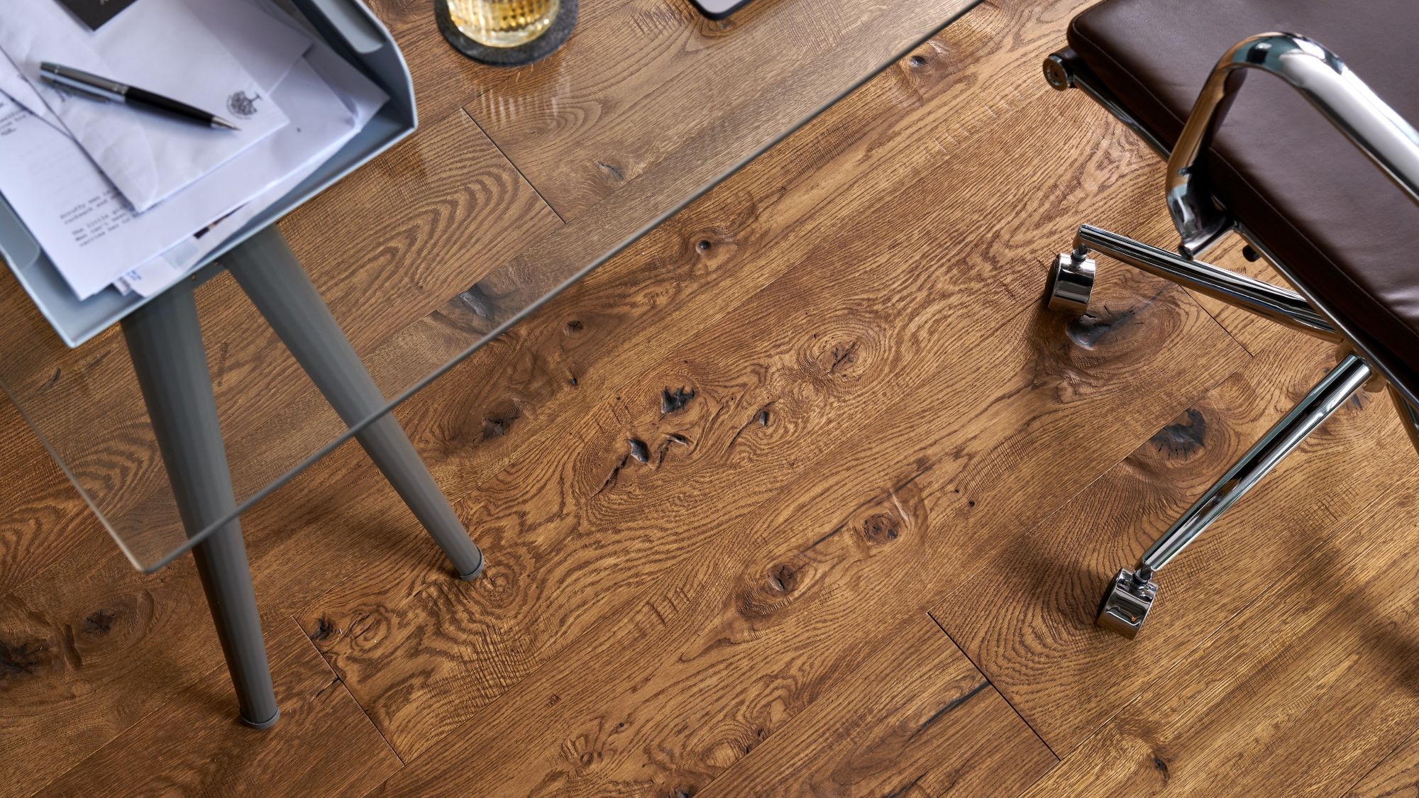 A top-down view of Berkeley Cathedral Oak engineered wood flooring featuring deep grain patterns and rustic knots, partially covered by a glass desk and a leather office chair.