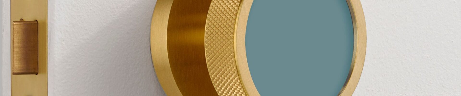 An extreme close-up of a modern, brushed gold brass mortice door knob mounted on a white door. The knob features a distinctive knurled texture around its edge for grip and a flat, circular face inset with a matte teal-blue colour.