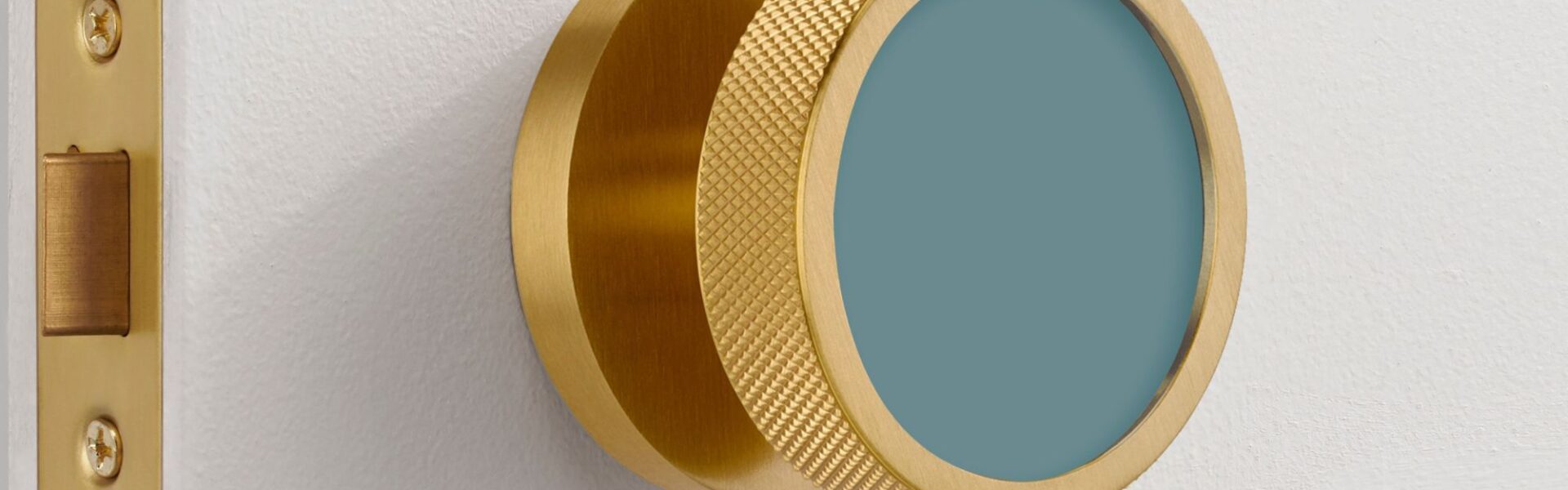 An extreme close-up of a modern, brushed gold brass mortice door knob mounted on a white door. The knob features a distinctive knurled texture around its edge for grip and a flat, circular face inset with a matte teal-blue colour.