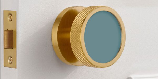 An extreme close-up of a modern, brushed gold brass mortice door knob mounted on a white door. The knob features a distinctive knurled texture around its edge for grip and a flat, circular face inset with a matte teal-blue colour.