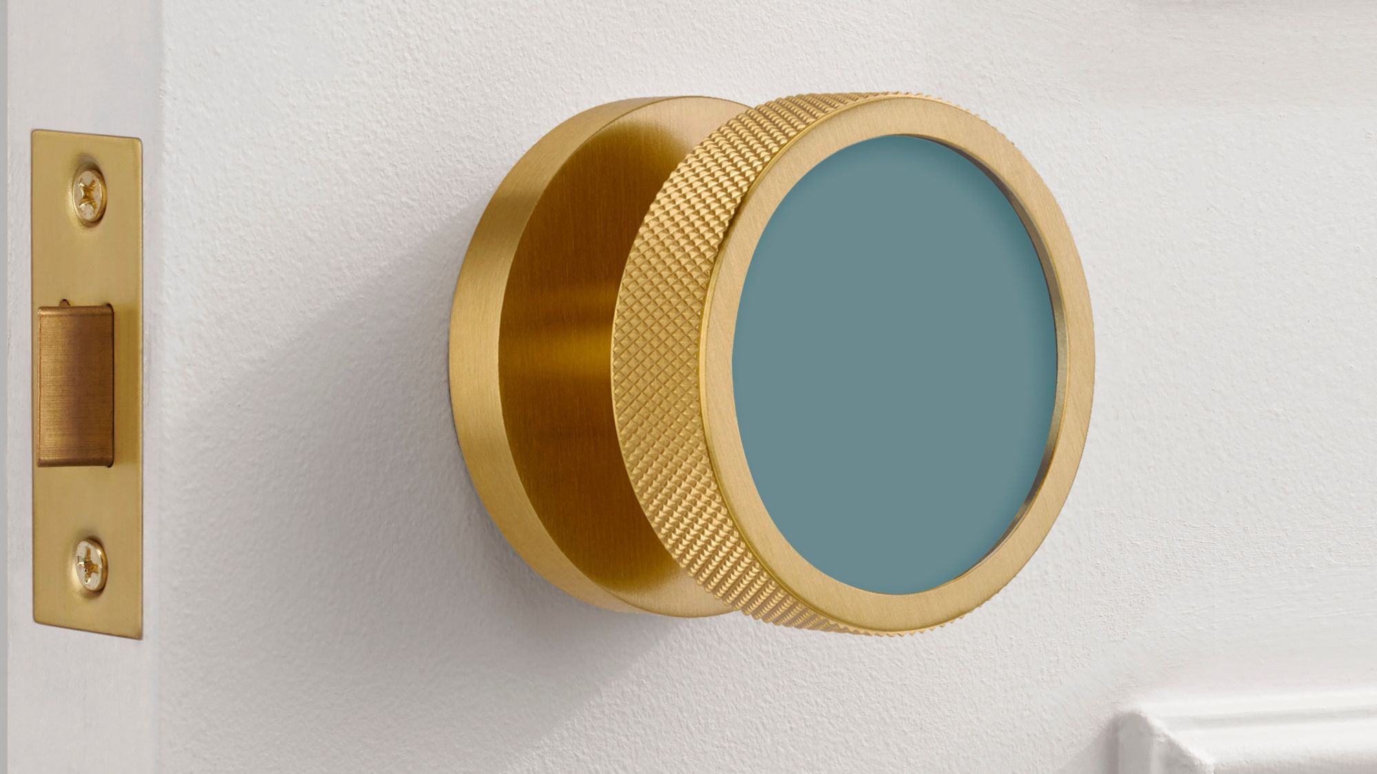 An extreme close-up of a modern, brushed gold brass mortice door knob mounted on a white door. The knob features a distinctive knurled texture around its edge for grip and a flat, circular face inset with a matte teal-blue colour.