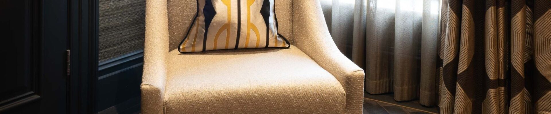 A luxury interior scene featuring an oatmeal-coloured textured armchair with a geometric gold and black silk cushion. The chair sits on dark, herringbone-patterned Charred Oak engineered wood flooring, positioned against a dark textured wall and charcoal grey door trim.