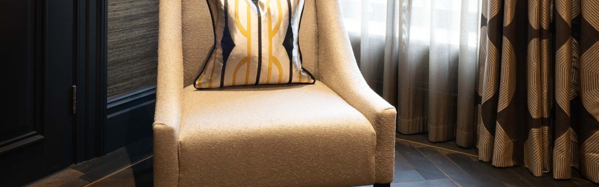 A luxury interior scene featuring an oatmeal-coloured textured armchair with a geometric gold and black silk cushion. The chair sits on dark, herringbone-patterned Charred Oak engineered wood flooring, positioned against a dark textured wall and charcoal grey door trim.