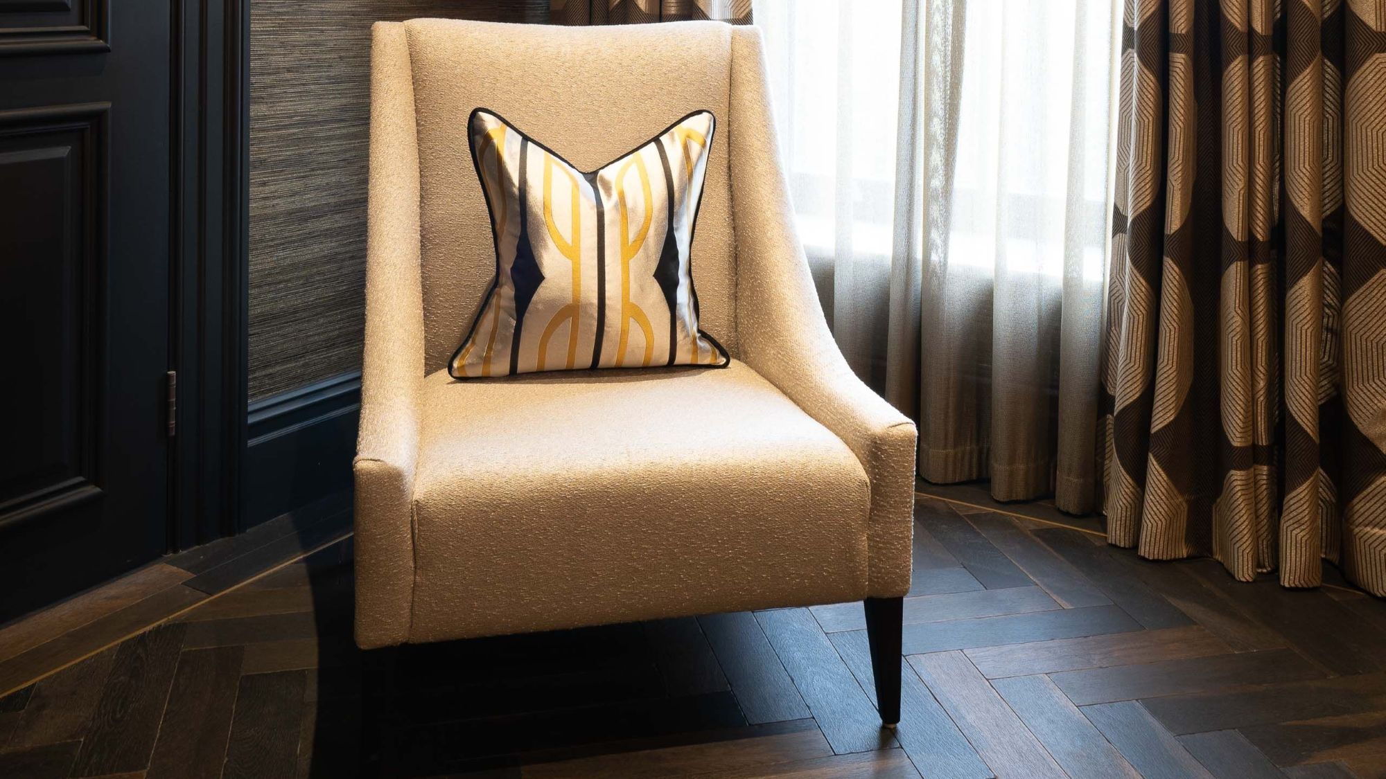 A luxury interior scene featuring an oatmeal-coloured textured armchair with a geometric gold and black silk cushion. The chair sits on dark, herringbone-patterned Charred Oak engineered wood flooring, positioned against a dark textured wall and charcoal grey door trim.