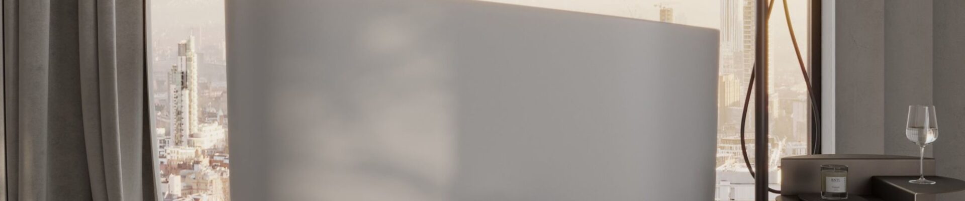 A minimalist, high-angle interior shot of a modern bathroom featuring a matte white stone Japanese soaking tub as the centrepiece. The tub has a sleek, asymmetrical rim that rises higher on one side, positioned in front of a floor-to-ceiling window overlooking a hazy city skyline at sunset.