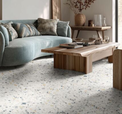 A modern living room featuring Venetica Terrazzo floor tiles with a white base and grey, blue, and gold chips. The room is styled with a curved sage green sofa, natural wood coffee tables and earthy organic decor.