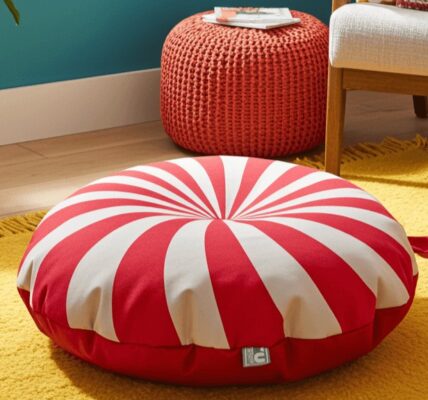 A red and white striped circular floor cushion sits on a textured yellow rug. In the background, there is a woven orange ottoman, a white armchair, and a wicker basket on a light wood floor against a teal wall.