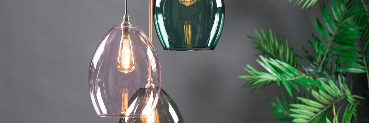 A three-light cluster chandelier featuring oval-shaped, translucent glass shades in teal, soft pink, and smoky grey. The pendants are suspended at staggered heights by fabric-covered cords against a dark grey background, with a green palm plant visible on the right.