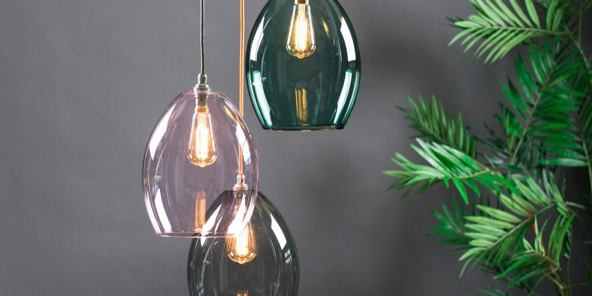 A three-light cluster chandelier featuring oval-shaped, translucent glass shades in teal, soft pink, and smoky grey. The pendants are suspended at staggered heights by fabric-covered cords against a dark grey background, with a green palm plant visible on the right.