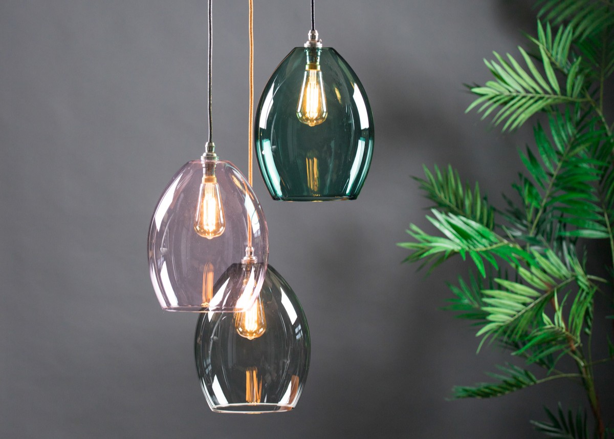 A three-light cluster chandelier featuring oval-shaped, translucent glass shades in teal, soft pink, and smoky grey. The pendants are suspended at staggered heights by fabric-covered cords against a dark grey background, with a green palm plant visible on the right.