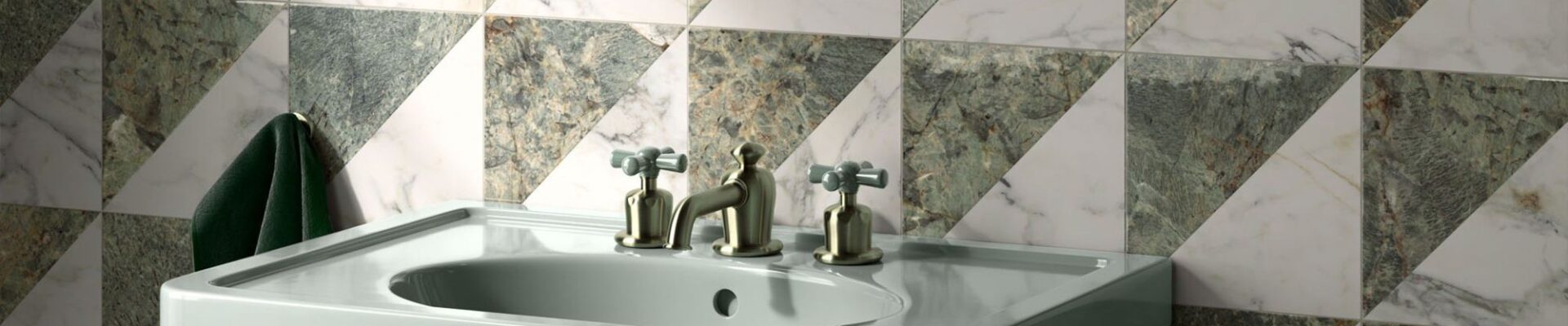 A high-angle, close-up shot of a sage green pedestal sink against a wall of geometric marble tiles. The square tiles are divided diagonally into two triangles: one of white marble with grey veining and the other of dark green marble with gold and white veining. The tiles are arranged in a repeating pattern of alternating triangles.