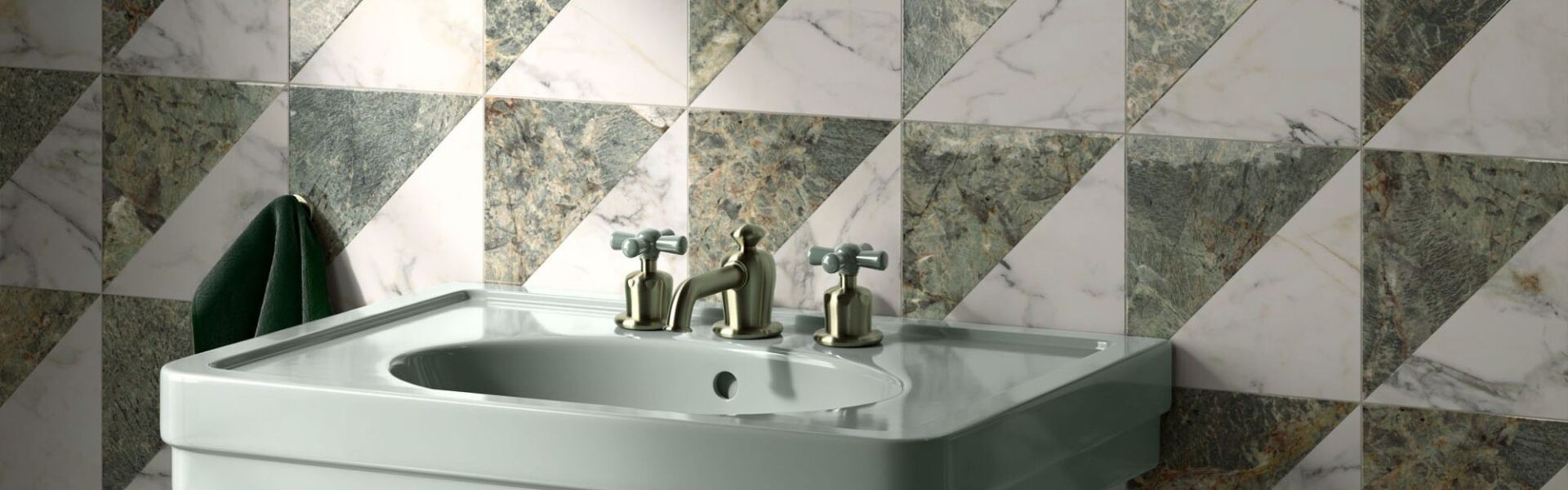 A high-angle, close-up shot of a sage green pedestal sink against a wall of geometric marble tiles. The square tiles are divided diagonally into two triangles: one of white marble with grey veining and the other of dark green marble with gold and white veining. The tiles are arranged in a repeating pattern of alternating triangles.