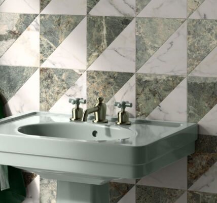A high-angle, close-up shot of a sage green pedestal sink against a wall of geometric marble tiles. The square tiles are divided diagonally into two triangles: one of white marble with grey veining and the other of dark green marble with gold and white veining. The tiles are arranged in a repeating pattern of alternating triangles.