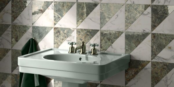 A high-angle, close-up shot of a sage green pedestal sink against a wall of geometric marble tiles. The square tiles are divided diagonally into two triangles: one of white marble with grey veining and the other of dark green marble with gold and white veining. The tiles are arranged in a repeating pattern of alternating triangles.