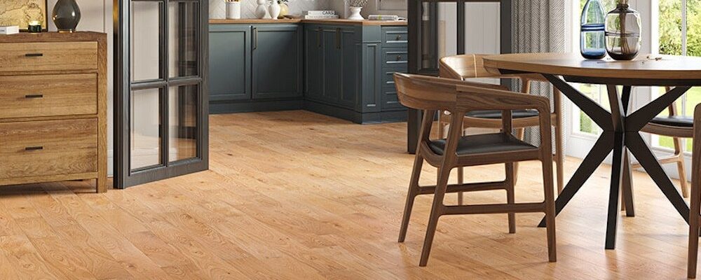 A modern open-plan kitchen and dining area featuring warm Lusso Carrara Orchard engineered oak flooring, dark teal cabinetry, and mid-century modern wooden furniture.