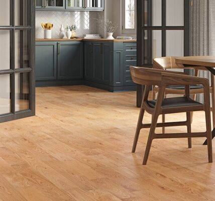 A modern open-plan kitchen and dining area featuring warm Lusso Carrara Orchard engineered oak flooring, dark teal cabinetry, and mid-century modern wooden furniture.