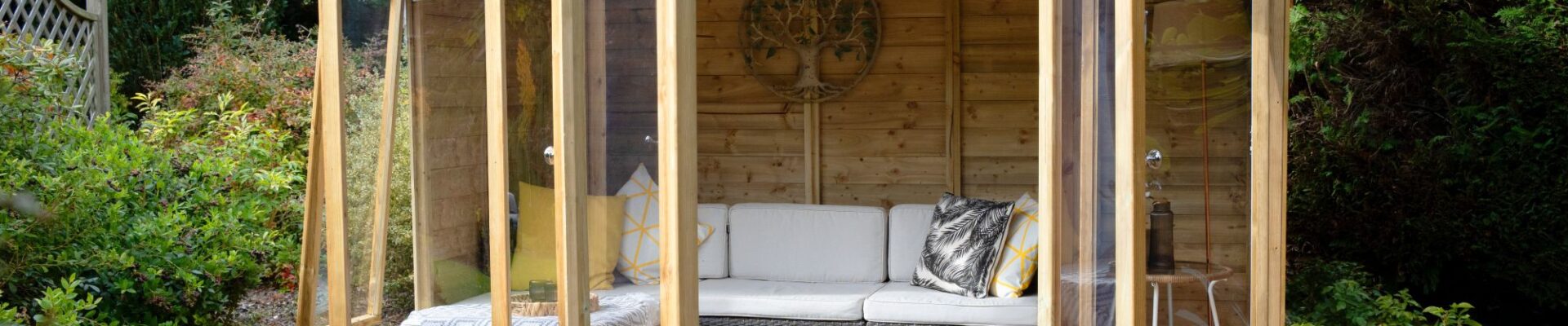 A wide-angle, eye-level shot shows a modern, light-wood Beckwood summerhouse nestled in a lush garden setting. The structure features double glass doors and large floor-to-ceiling windows, all of which are swung open to reveal an inviting interior.