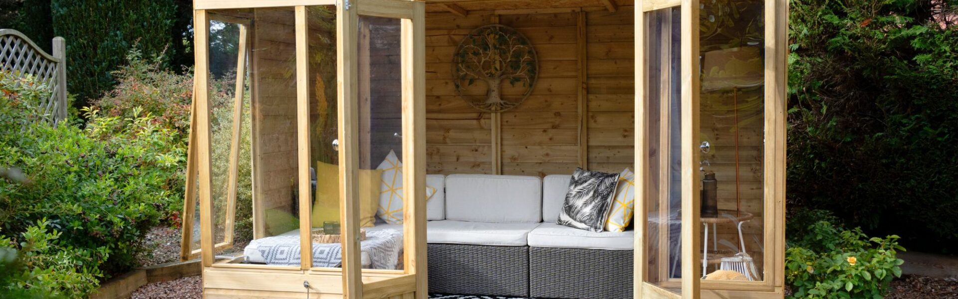 A wide-angle, eye-level shot shows a modern, light-wood Beckwood summerhouse nestled in a lush garden setting. The structure features double glass doors and large floor-to-ceiling windows, all of which are swung open to reveal an inviting interior.