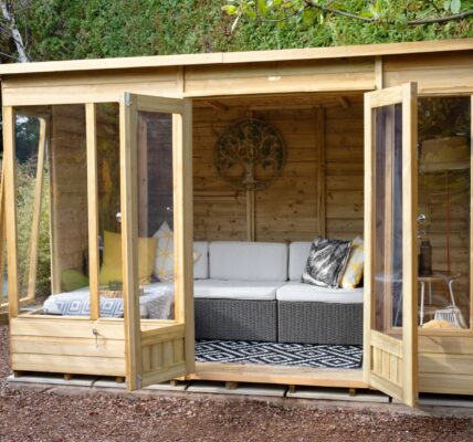 A wide-angle, eye-level shot shows a modern, light-wood Beckwood summerhouse nestled in a lush garden setting. The structure features double glass doors and large floor-to-ceiling windows, all of which are swung open to reveal an inviting interior.