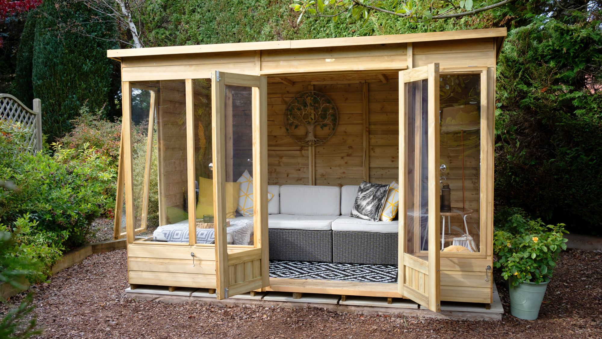 A wide-angle, eye-level shot shows a modern, light-wood Beckwood summerhouse nestled in a lush garden setting. The structure features double glass doors and large floor-to-ceiling windows, all of which are swung open to reveal an inviting interior.