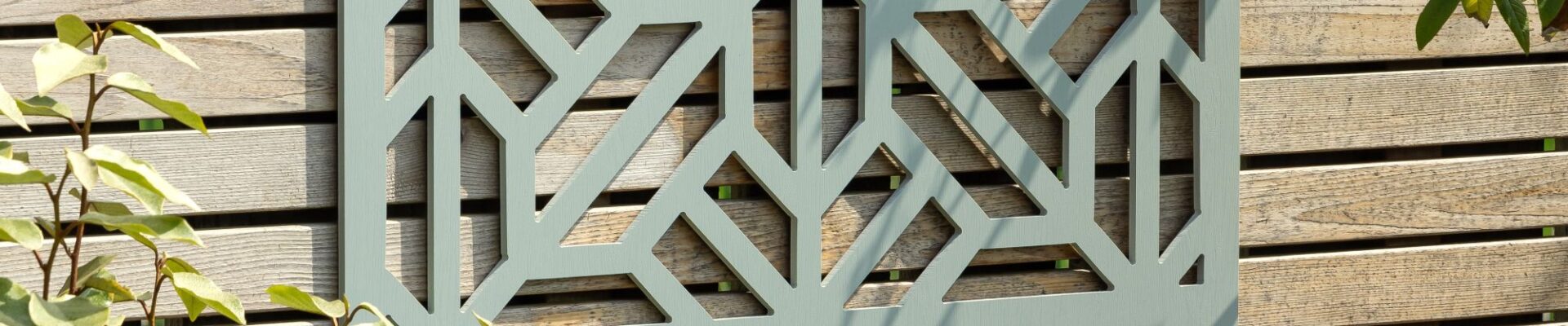 A decorative, square, sage-green wooden privacy screen with an intricate geometric, star-shaped cut-out pattern is mounted on a light-coloured, slatted wooden fence. The screen is centred in the upper portion of the frame, showcasing the detailed symmetry of the cutouts against the fence's wood grain.
