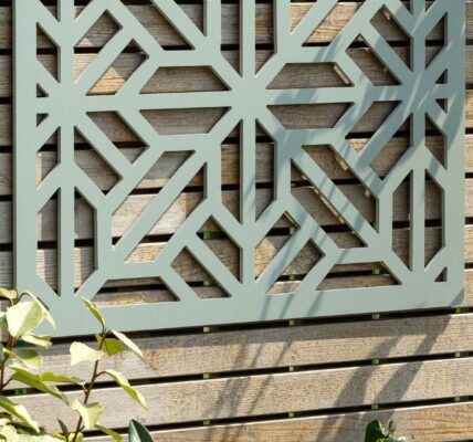 A decorative, square, sage-green wooden privacy screen with an intricate geometric, star-shaped cut-out pattern is mounted on a light-coloured, slatted wooden fence. The screen is centred in the upper portion of the frame, showcasing the detailed symmetry of the cutouts against the fence's wood grain.