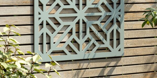 A decorative, square, sage-green wooden privacy screen with an intricate geometric, star-shaped cut-out pattern is mounted on a light-coloured, slatted wooden fence. The screen is centred in the upper portion of the frame, showcasing the detailed symmetry of the cutouts against the fence's wood grain.