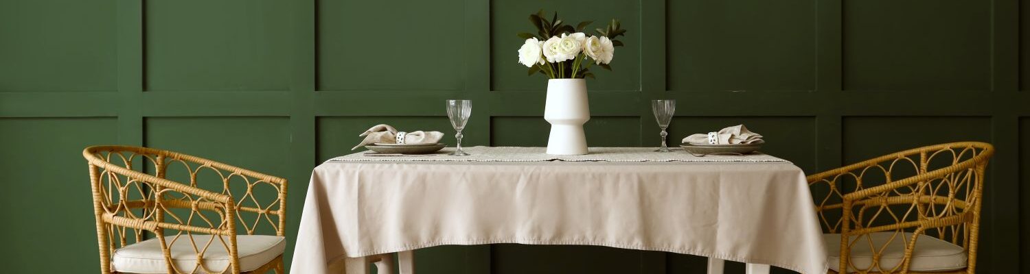 An elegant, eye-level wide shot of a dining area featuring a deep pepper tree green wainscoted wall. Centred in the room is a rectangular white wooden dining table draped with a light beige tablecloth, set with two minimalist place settings, crystal wine glasses, and a white vase holding white ranunculus flowers.